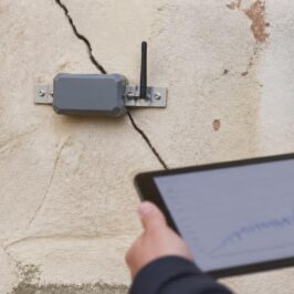 Sensore wireless per il monitoraggio delle fessure installato su una lesione diagonale di una parete intonacata, con tecnico che consulta i dati su tablet
